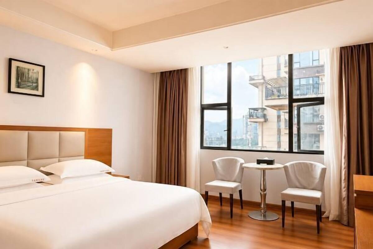 Een hotelkamer met een groot bed, twee stoelen, een kleine tafel en een groot raam met uitzicht op de stad.