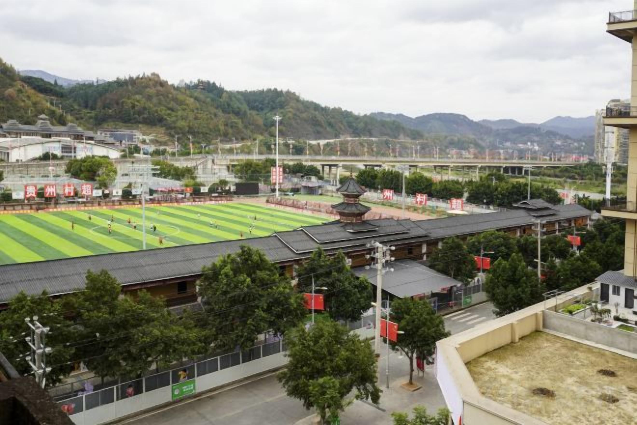A sports field with a traditional pavilion, surrounded by buildings and trees.