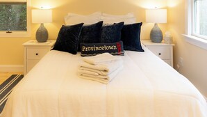 A neatly made bed with white linens, a folded towel, and a pillow with "Provincetown" embroidered on it. There are two bedside lamps and a baseball bat hanging on the wall. - Charming studio in delightful Provincetown with WiFi, AC (Provincetown)
