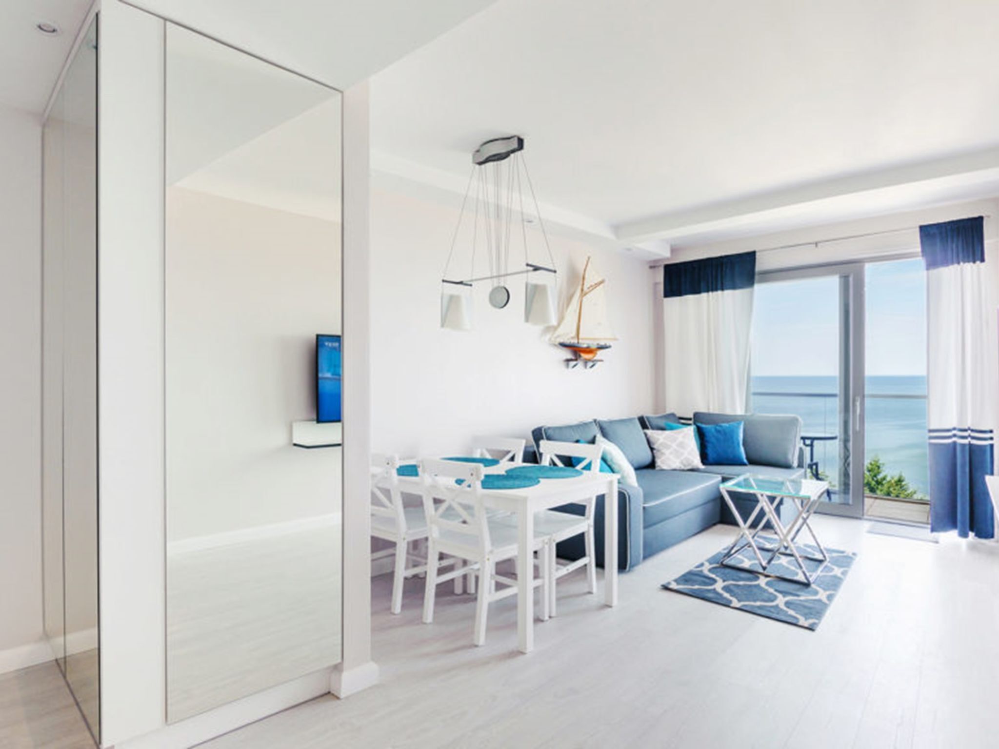 Een moderne woonkamer met een bank, eettafel, stoelen en uitzicht op zee door grote ramen.