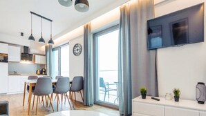 Een moderne woonkamer met een ronde witte tafel, grijze stoelen, een witte bank, een aan de muur bevestigde televisie, en een balkon met gordijnen.