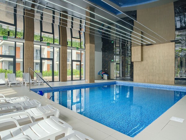 An indoor swimming pool with clear blue water, white lounge chairs, and a modern interior design with large windows and a view of greenery outside.