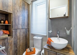 A bathroom with a wooden cabinet, a toilet, a sink with a mirror above, and a window with blinds. - Luxurious Lodge, perfect for couples or familes, sleeping four people. (Nr Kintore)