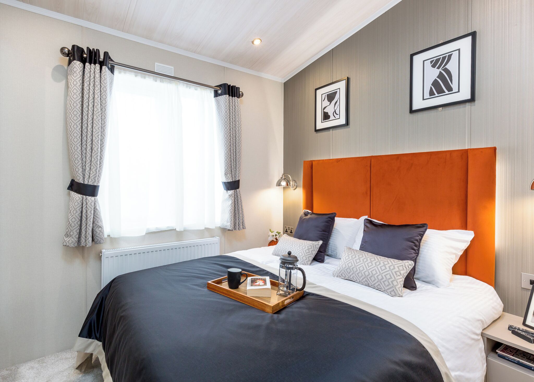 A hotel room with a large bed, a bedside table with a lamp, a tray with a mug and a book, and two framed pictures on the wall.