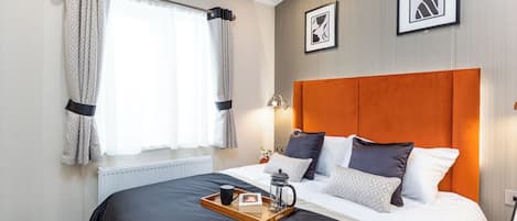 A hotel room with a large bed, a bedside table with a lamp, a tray with a mug and a book, and two framed pictures on the wall.