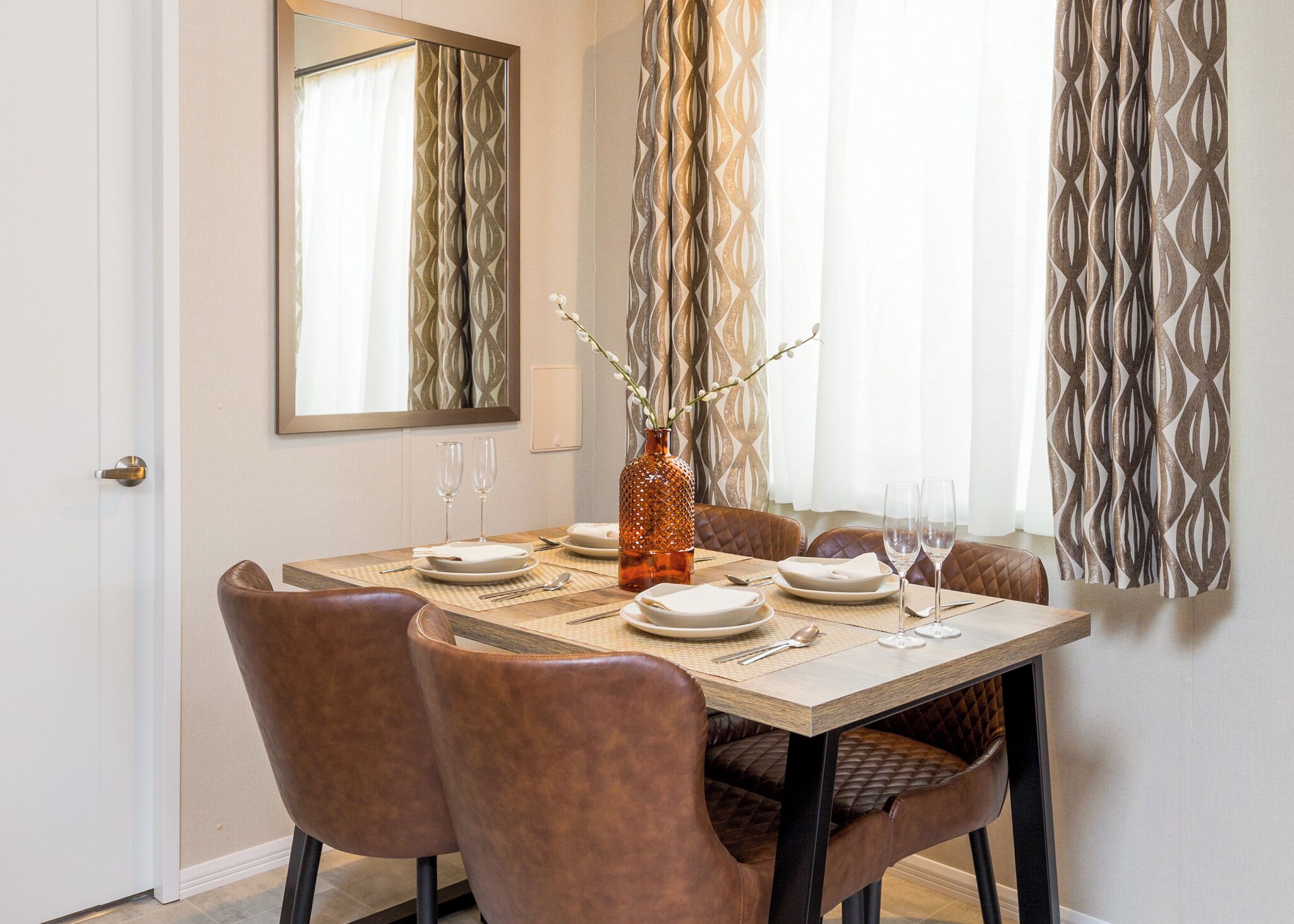 Un comedor con una mesa dispuesta para seis, un florero con ramas y un espejo en la pared.