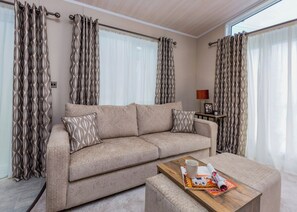 A modern living room with a beige sofa, patterned cushions, a wooden coffee table with magazines, and a lamp.