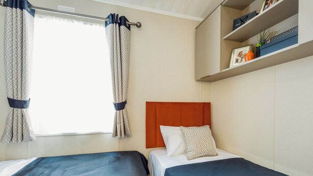 A hotel room with two beds, a window with curtains, a shelf with decorative items, and a headboard.