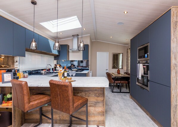 A modern kitchen with a central island, bar stools, a dining area, and built-in appliances. - Luxurious Lodge, perfect for couples or familes, sleeping four people. (Nr Kintore)