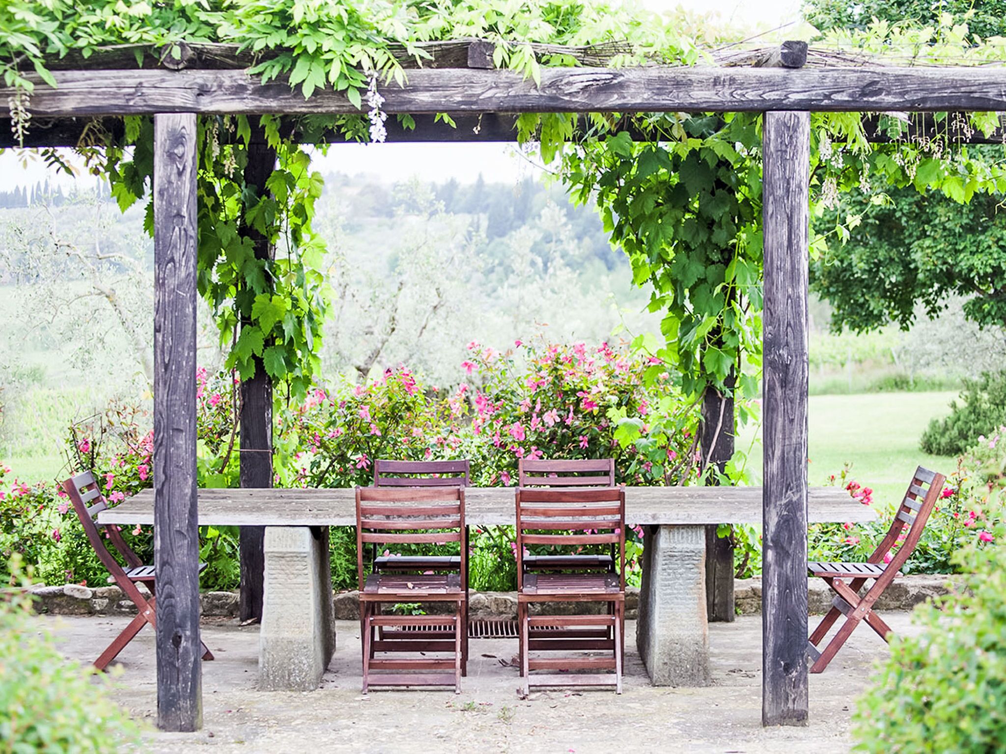A wooden pergola with two wooden benches under it, surrounded by lush greenery and pink flowers.
