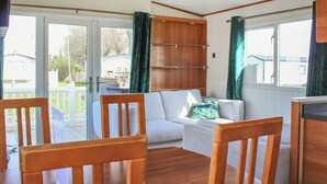 Una habitación con piso de madera, sofá blanco, sillas de madera, una mesita, un armario de madera y un ventanal corredizo que da a un espacio exterior.