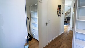A hallway with white walls, wooden flooring, and a white door with a keyhole. There are white shelves on the left and a small table with a chair in the background. - Dat Huus by Interhome (Zinnowitz)