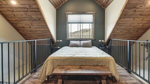 Una camera da letto con soffitto spiovente, un letto con copripiumino beige, una finestra con persiane e una panca in legno.
