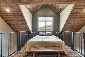 A bedroom with a sloped ceiling, a bed with a beige bedspread, a window with blinds, and a wooden bench.