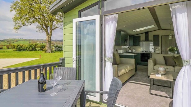 A balcony with a table and chairs, a sliding glass door leading to a living area with a sofa and a small table, and a view of a green landscape. - 2 bedroom accommodation in Caersws (Caersws)