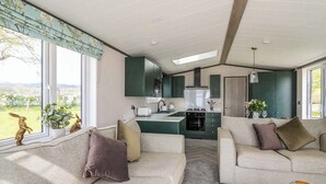 A modern living room with a sofa and a coffee table, a kitchen with green cabinets, and a view of the outdoors. - 2 bedroom accommodation in Caersws (Caersws)