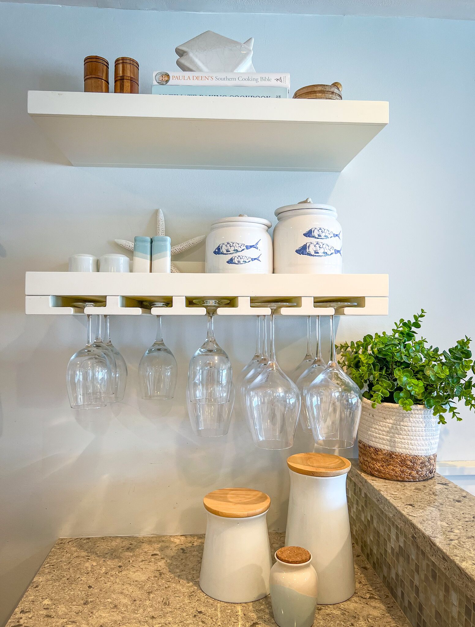 A kitchen counter with a white shelf holding a cookbook, salt shakers, and a decorative item. Below, there are jars with fish designs, a plant, and a wooden stool with a cork.