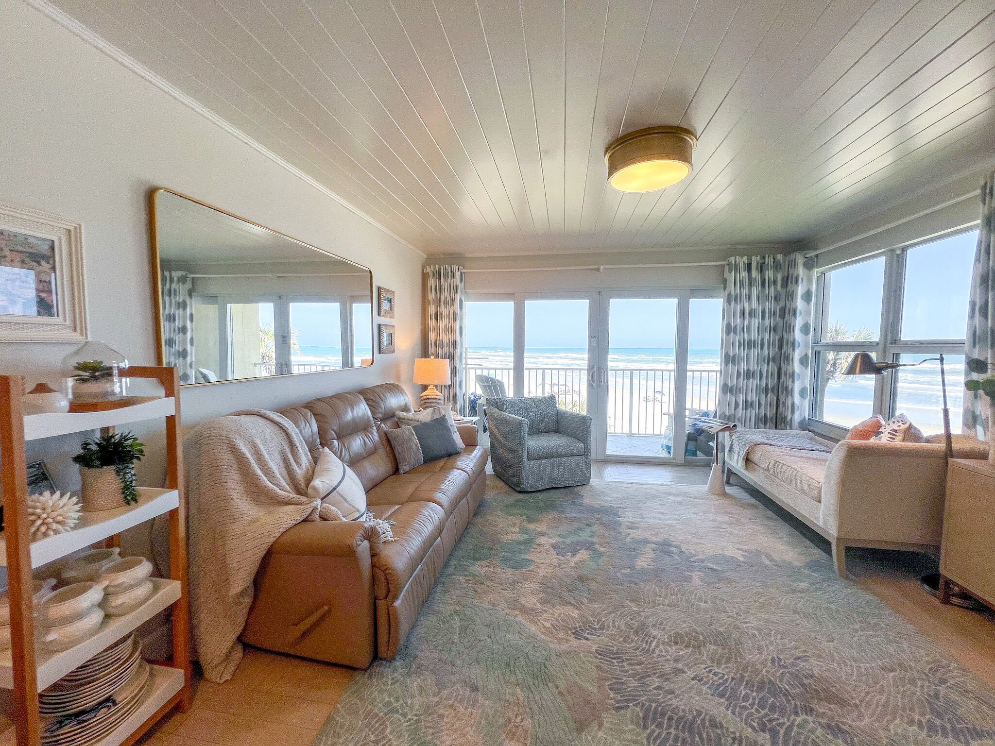 A spacious living room with a large sofa, a smaller armchair, and a dining area with a wooden table and chairs. A large window offers a view of the beach.