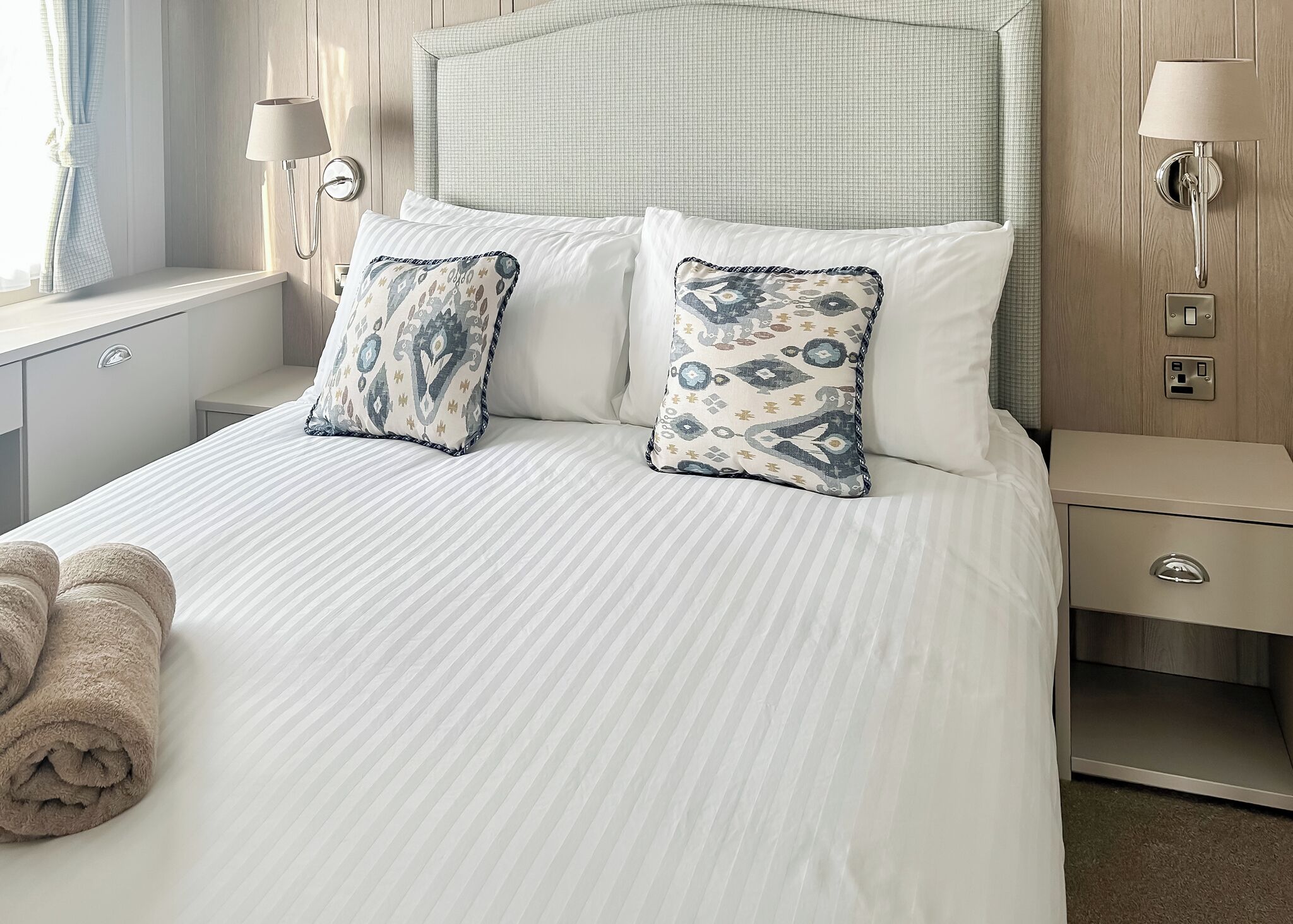 Una cama bien hecha con dos almohadones estampados, una mesita de noche con una l ámpara y un aplique en la pared.