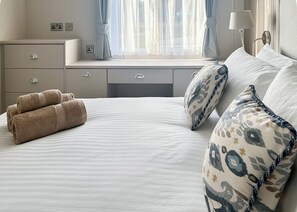 Cama bem arrumada com toalhas enroladas, mesa de cabeceira, abajur e janela com cortinas.