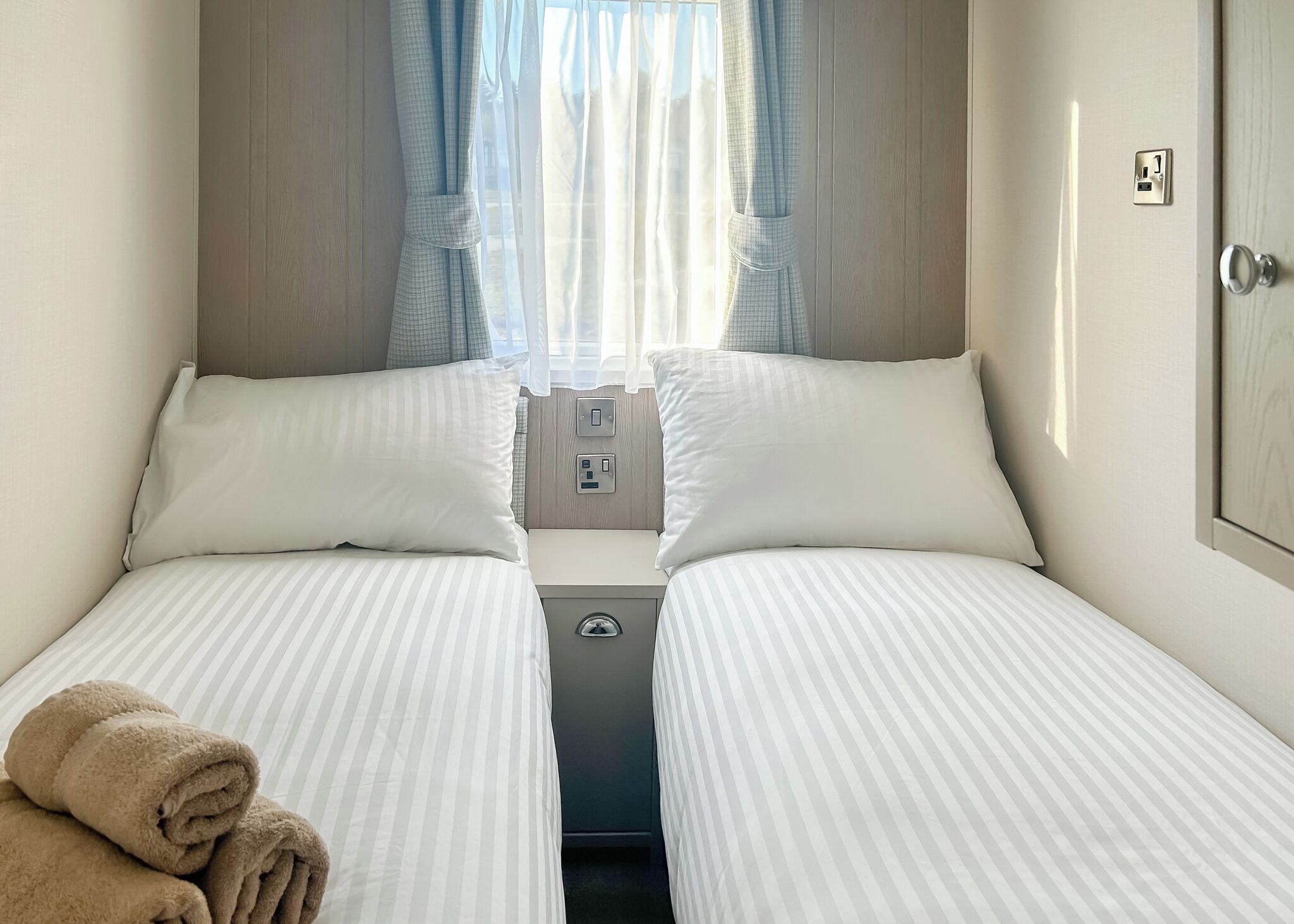 A twin room with two beds, a window with curtains, a bedside table, and a wall socket.