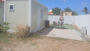 Ein kleines, eingeschossiges Gebäude mit einer Tür und einem Fenster, eine Terrasse mit Tisch und Stühlen sowie ein kleiner Garten.