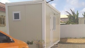 Ein kleines, beigefarbenes Einfamilienhaus mit Tür und Fenster, gelegen in einer Wohngegend. Im Vordergrund ein geparktes Auto und darüber eine Straßenlaterne.