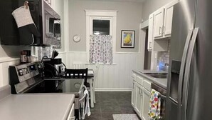 A compact kitchen with a stainless steel refrigerator, a built-in oven, and a microwave. There is a dining table with chairs and a window with patterned curtains.