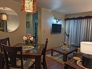 Zona pranzo con tavolo in vetro, sedie di legno, un vaso di fiori, una TV a schermo piatto montata a parete e un'ampia finestra con tende.