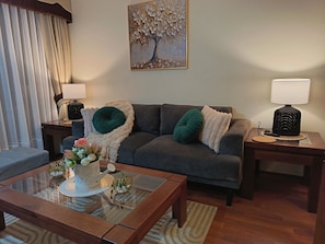 A living room with a sofa, two side tables with lamps, a glass coffee table with decorative items, and a painting on the wall.