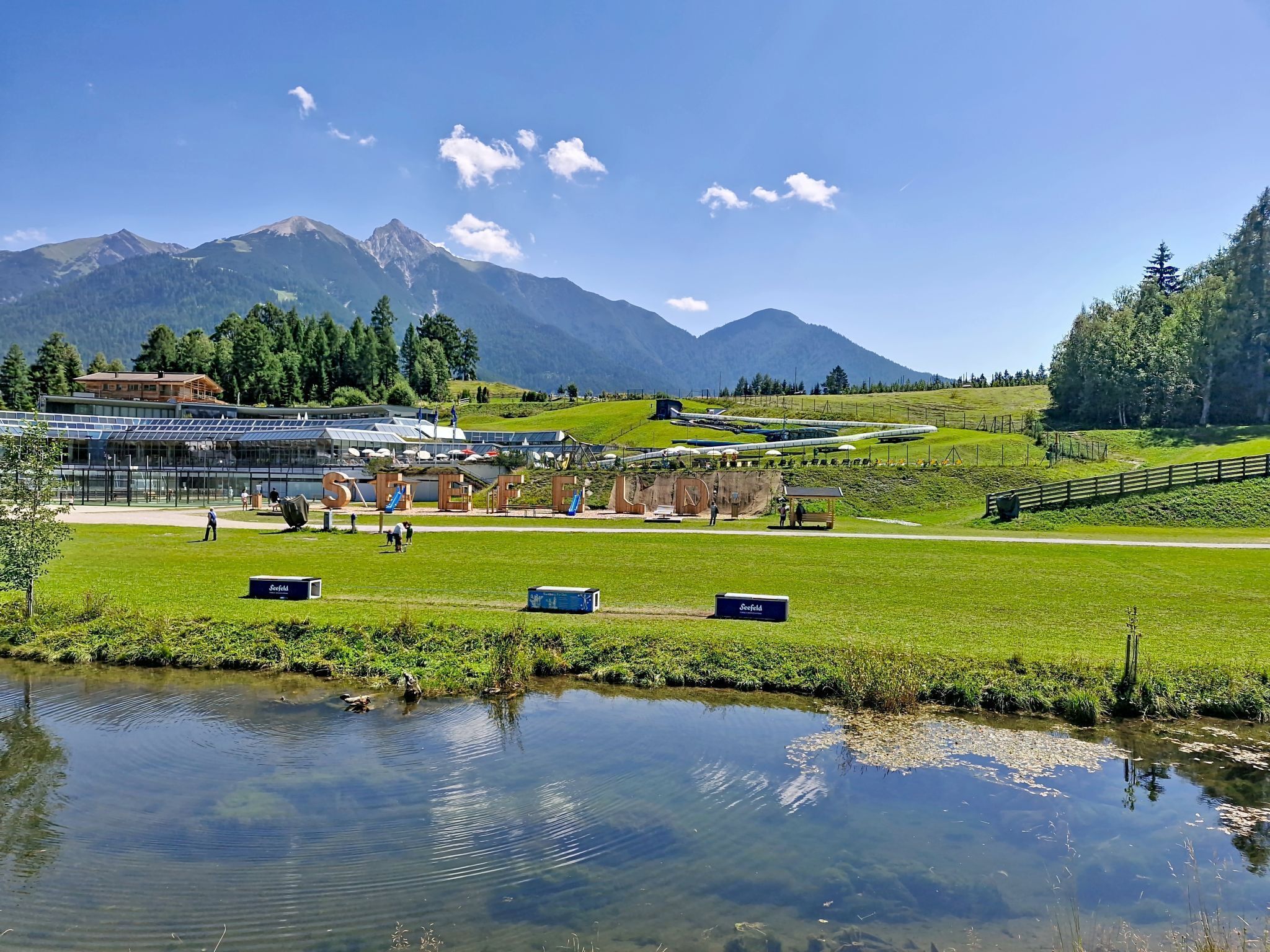 Vista de un lago con un edificio que tiene paneles solares, un área verde y montañas al fondo.
