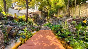 A garden pathway leading to a building with a modern design, surrounded by lush greenery and a small waterfall.