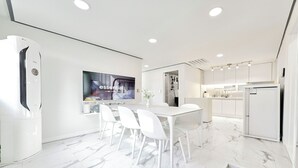 A modern kitchen with white cabinetry, a dining area with white chairs, and a large flat-screen TV mounted on the wall.