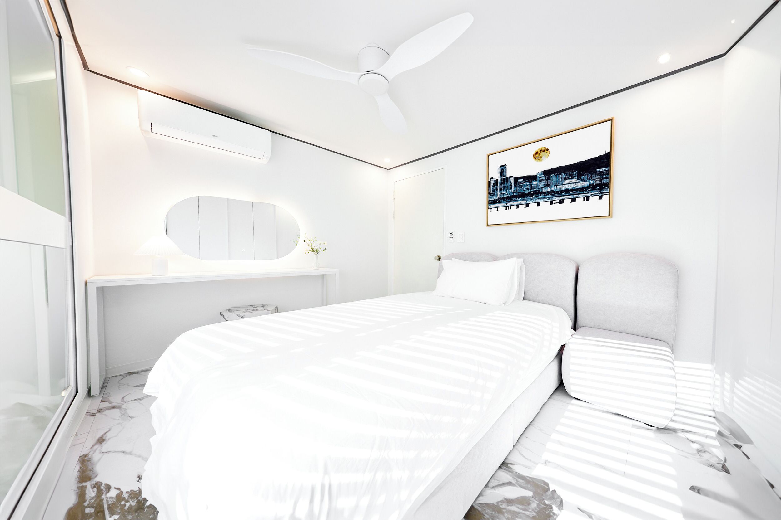 Quarto moderno com uma cama grande, um ventilador de teto, uma fotografia emoldurada de paisagem urbana e uma toucador branca com espelho.