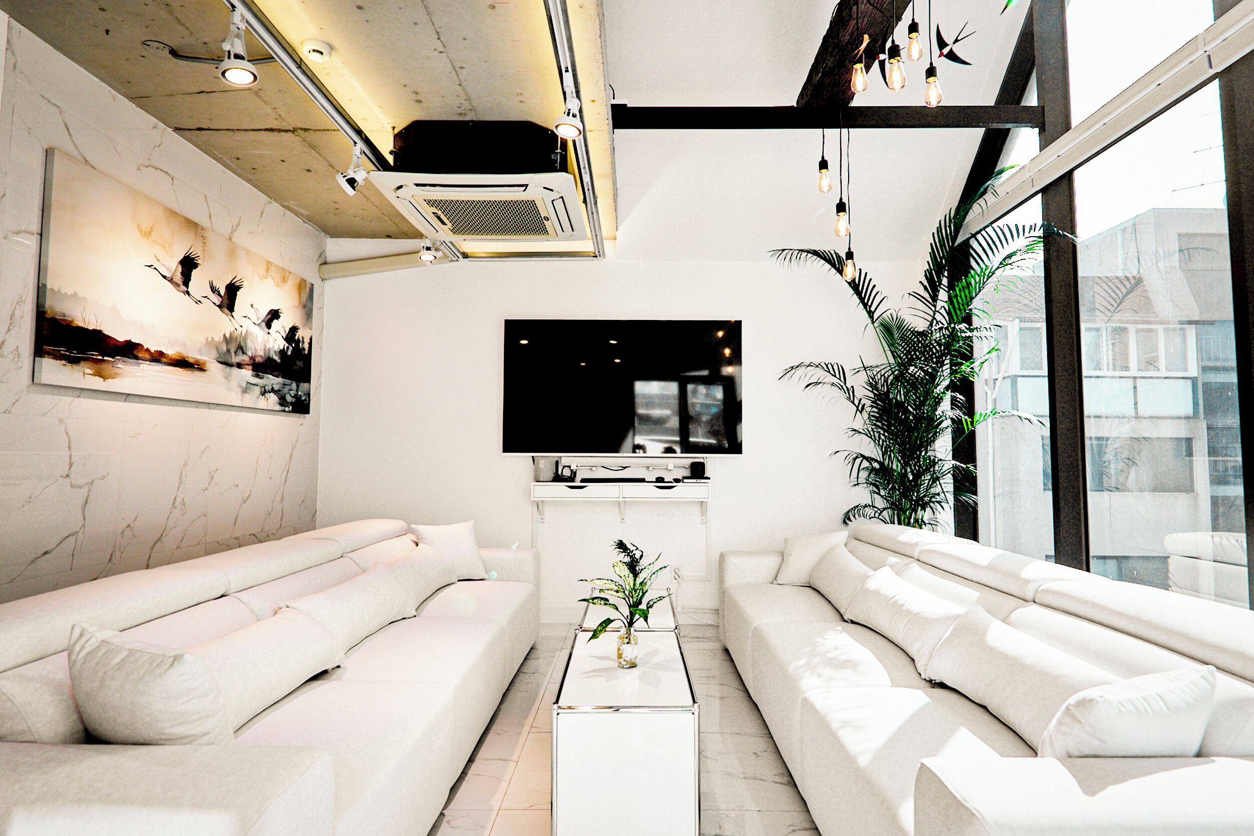A modern living room with white sofas, a flat-screen TV, and a large window providing a view of the city.