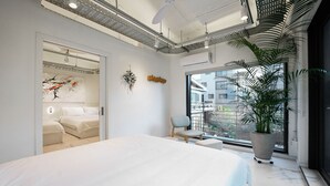 A modern bedroom with a large bed, a chair, a potted plant, and a view of the city through large windows.