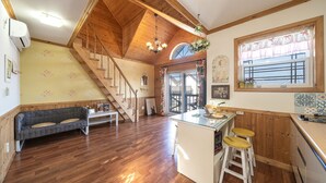 Un salón amplio con escalera de madera, un sofá, una mesa de comedor y una zona de cocina con taburetes.