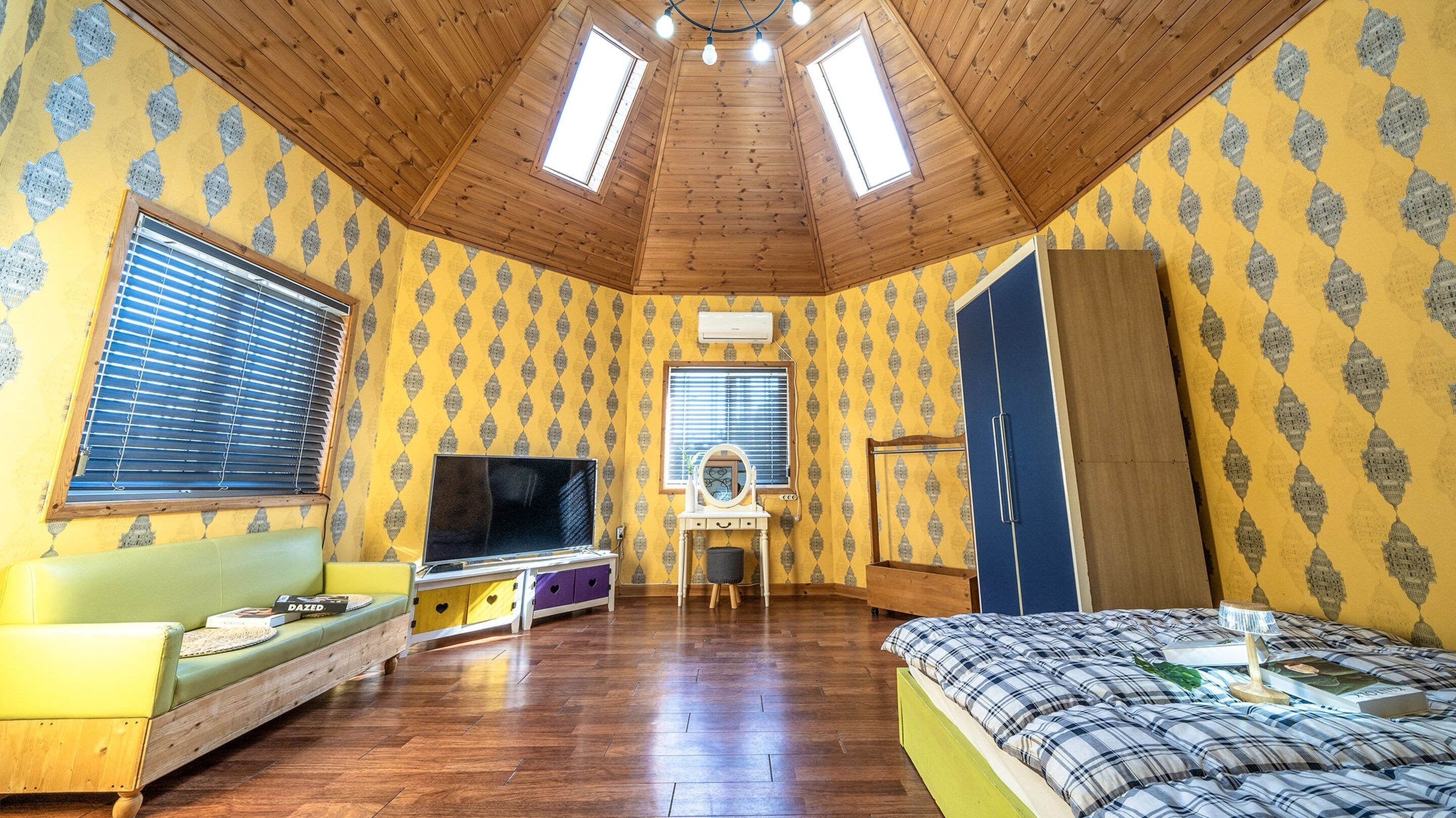 A room with a sloped ceiling, wooden beams, a bed with a plaid duvet, a TV, a vanity with a mirror, and a wardrobe.
