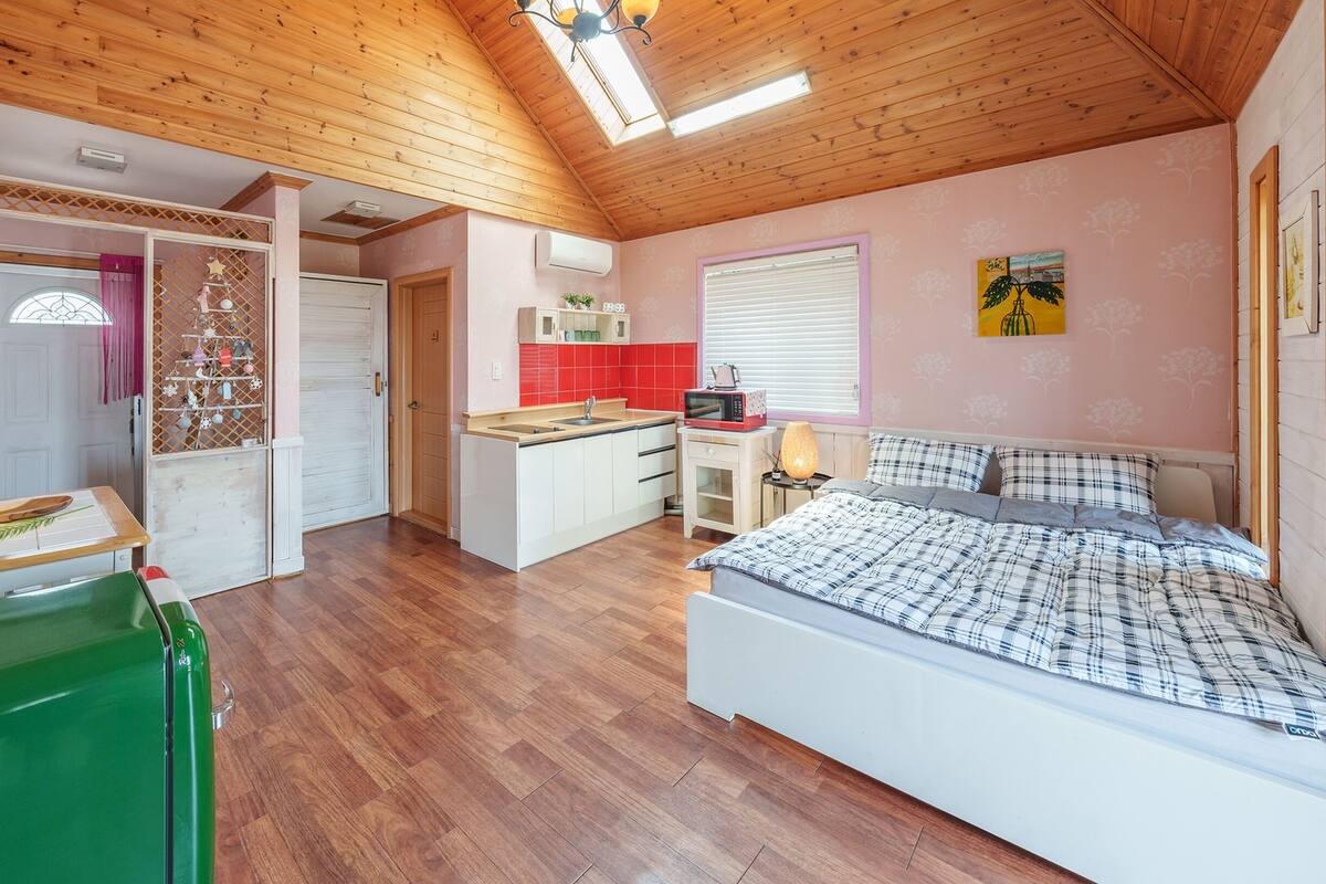 Un dormitorio con una cama, un refrigerador verde, una zona de cocina con un lavamanos y una ventana con persianas.