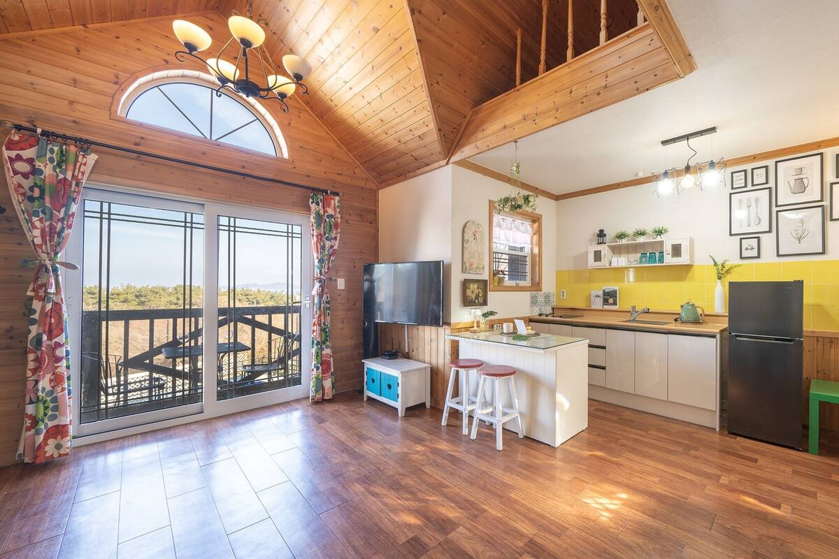 Una cocina amplia con suelo de madera, una isla grande con taburetes, un refrigerador empotrado y vistas a un espacio exterior por una puerta de vidrio.
