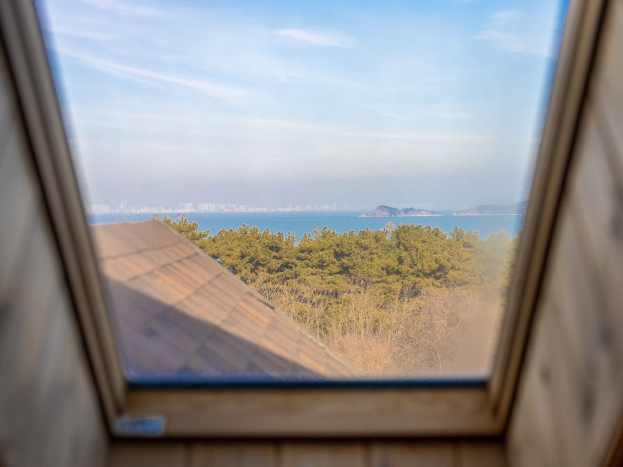 A view through a window with a wooden frame, overlooking a coastal landscape with trees and a distant city skyline.