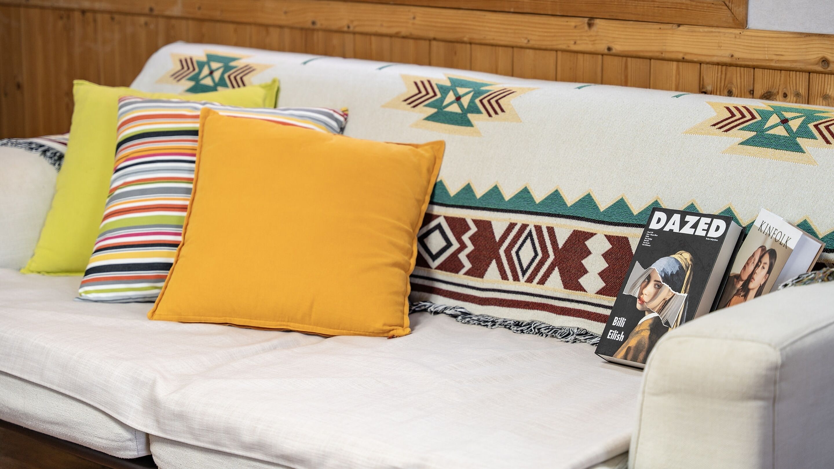 A sofa with a patterned blanket, a yellow pillow, and a magazine titled 'DAZED'.