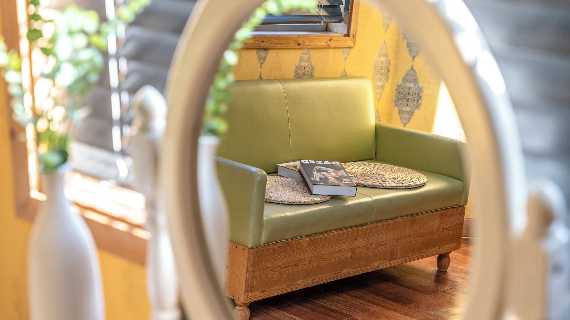 Un canapé en cuir vert, agrémenté d’un coussin tissé et d’un magazine, est placé dans une pièce dont le sol est en bois et un des murs est décoré d’un motif jaune.