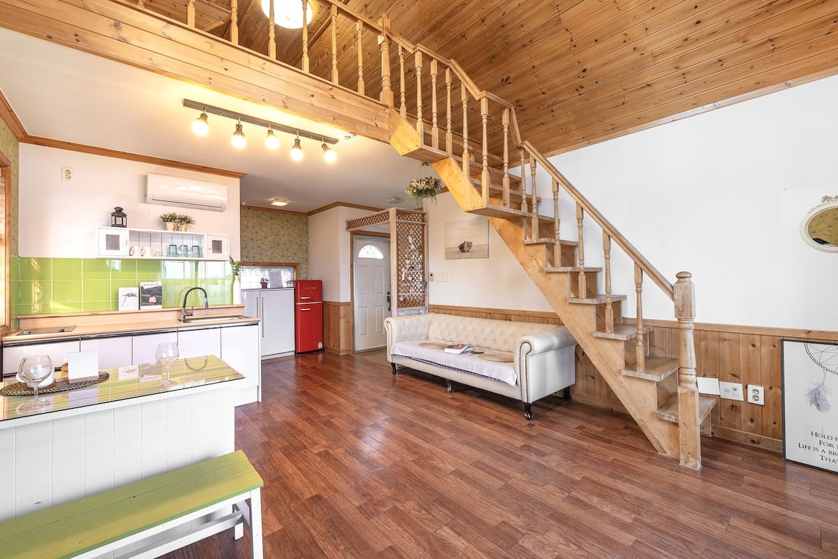 Un amplio salón con una escalera de madera, una cocina con azulejos verdes y un sofá blanco.