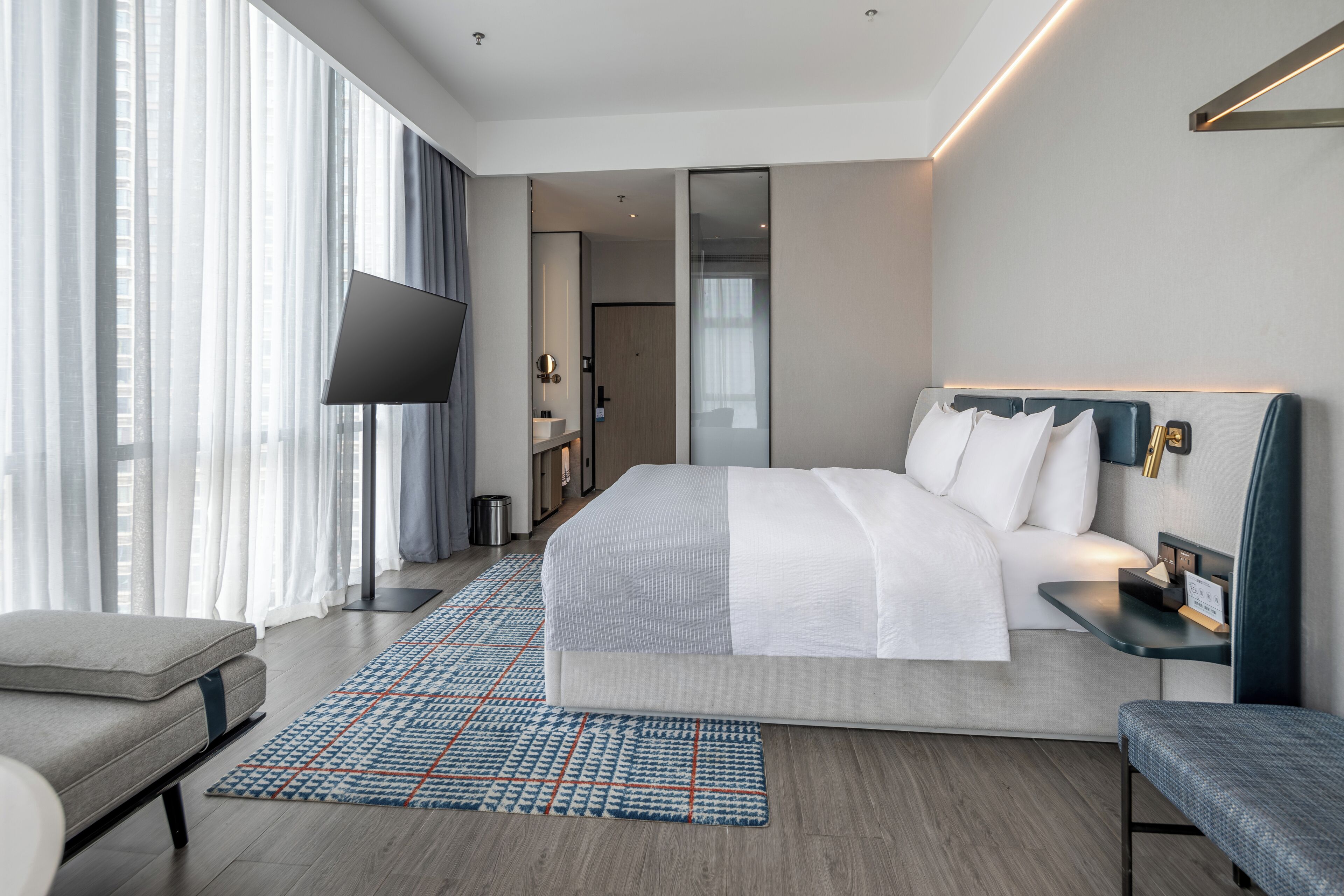 Ein modernes Hotelzimmer mit einem großen Bett, einem Nachttisch, einem Sessel, einem Fernseher und Blick auf die Stadt durch große Fenster.