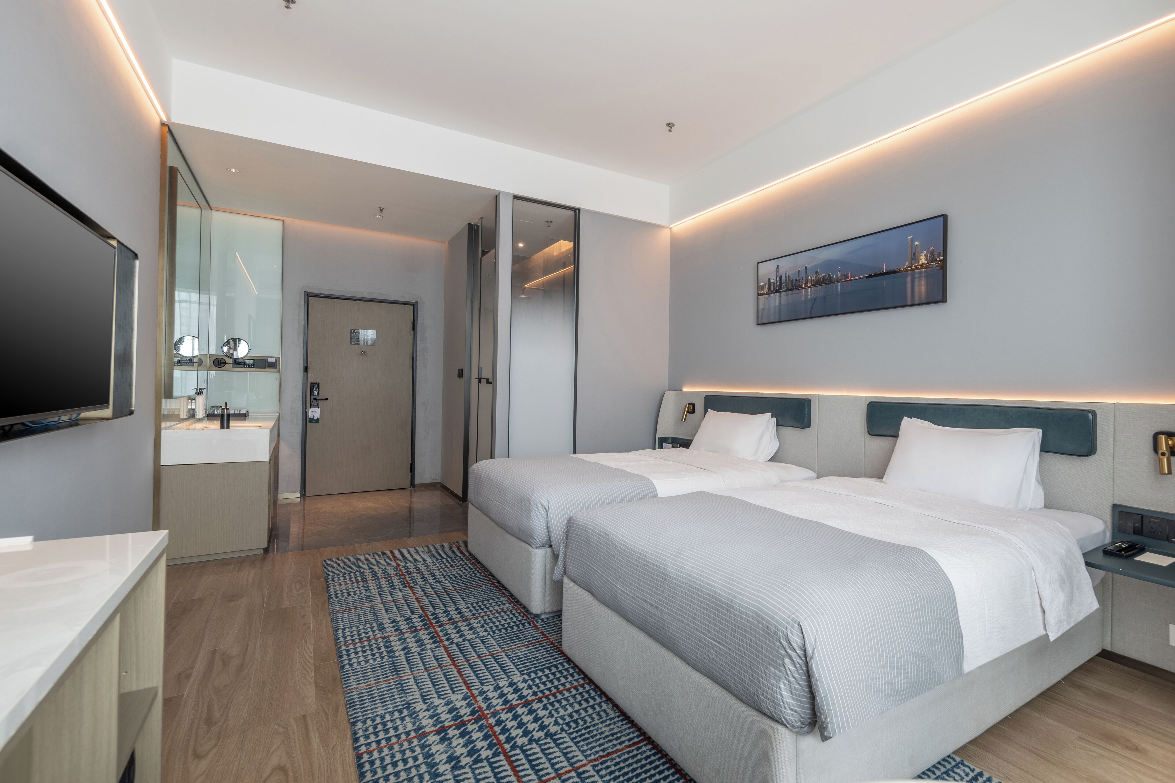 A modern hotel room with two beds, a flat-screen TV, a bathroom with a mirror, and a cityscape painting on the wall.