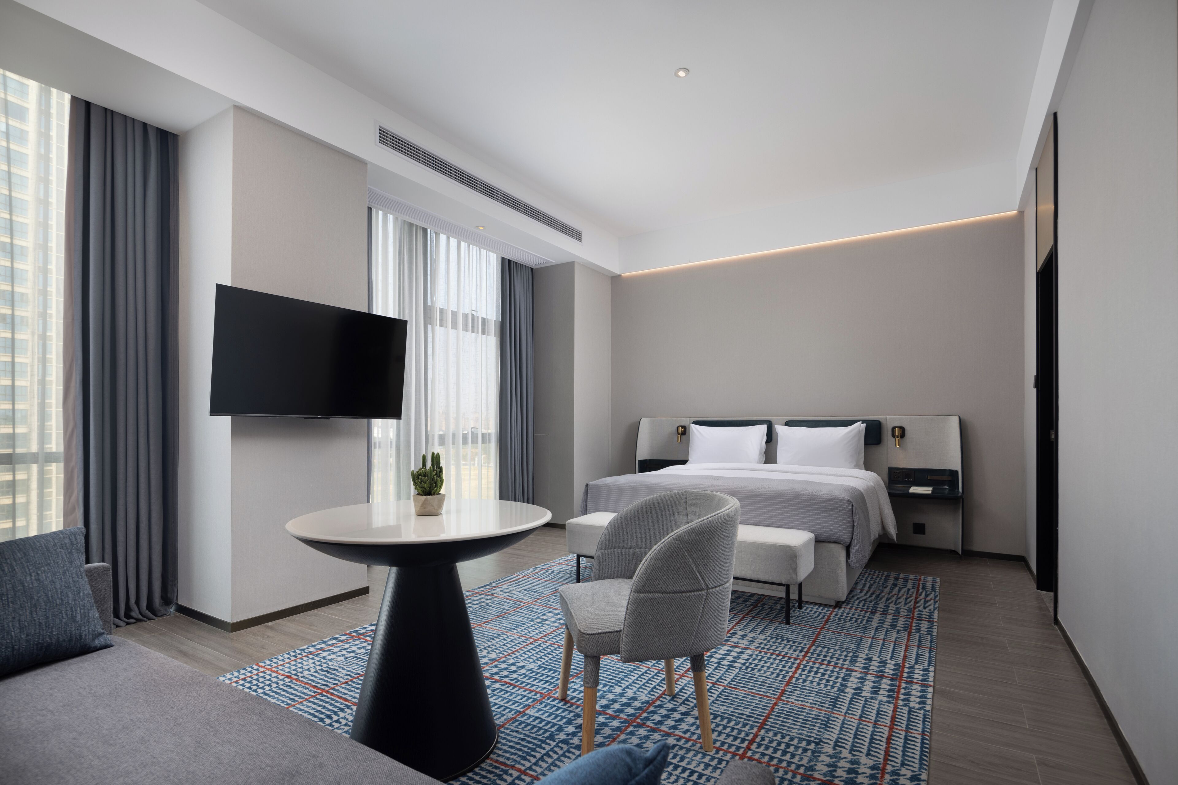 Una habitación de hotel moderna con una cama grande, un televisor de pantalla plana montado en la pared, una mesita redonda pequeña, un sillón gris y una alfombra estampada.