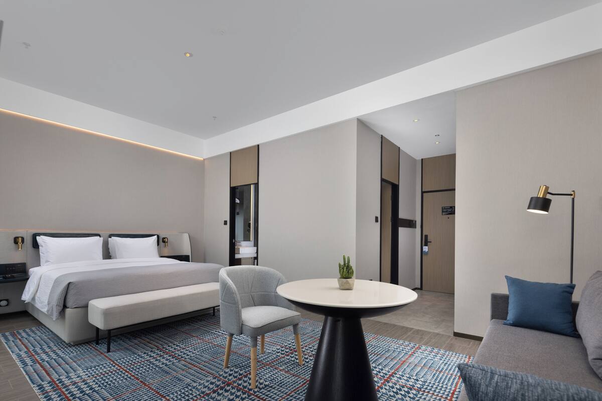 Moderni hotellihuone, jossa on sänky, oleskelualue, pieni pöytä ja lamppu.