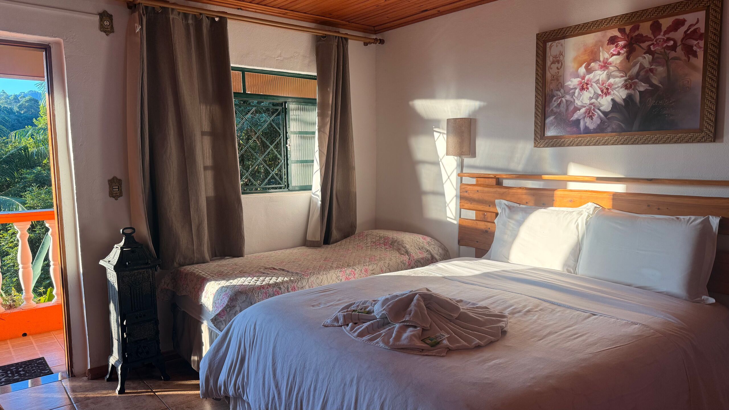 A hotel room with two beds, a large window with curtains, a painting of flowers, and a view of greenery outside.