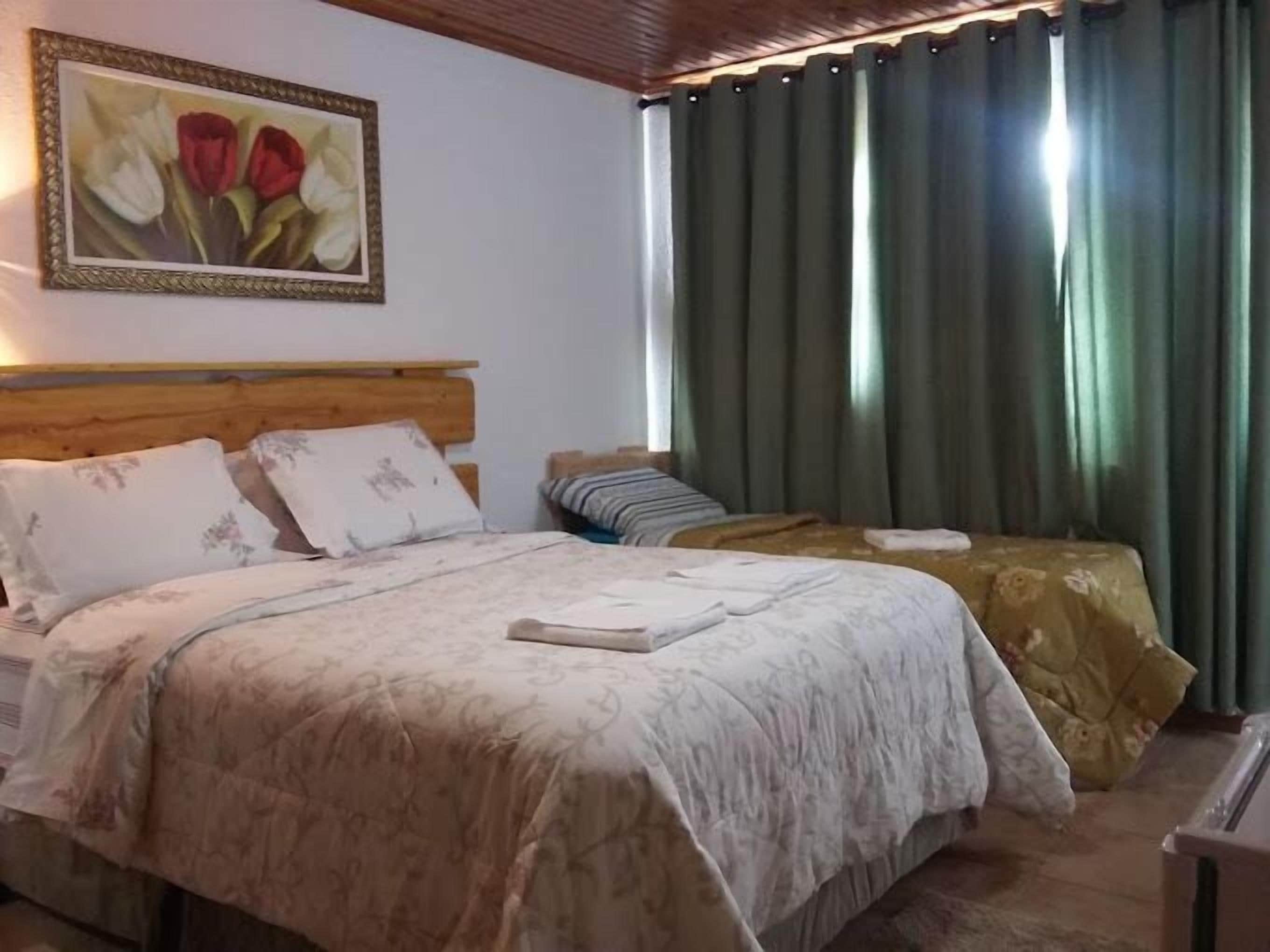 Quarto com cabeceira de madeira, duas camas com roupa de cama floral, mesa de cabeceira, mesa lateral, quadro decorativo com flores e cortinas verdes.